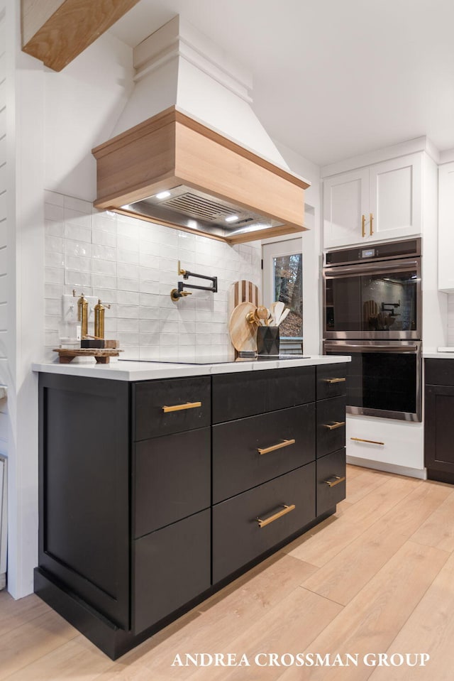 kitchen with tasteful backsplash, custom range hood, stainless steel double oven, light hardwood / wood-style floors, and white cabinetry