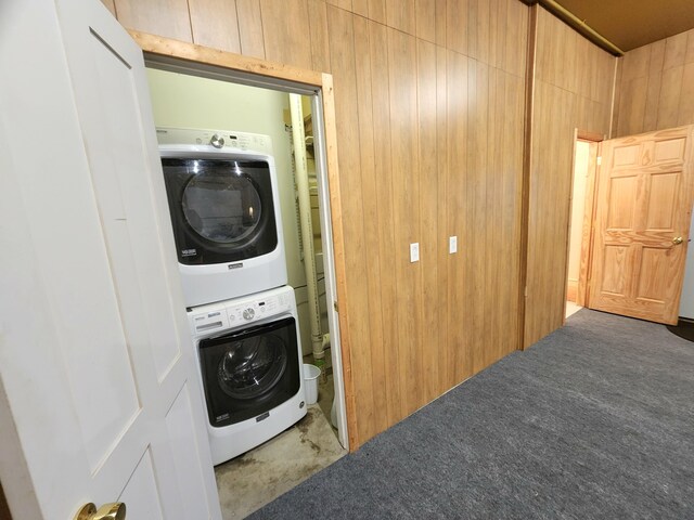 clothes washing area featuring dark colored carpet, wooden walls, and stacked washer and clothes dryer