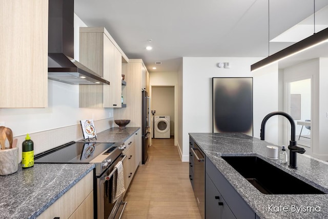 kitchen featuring dark stone counters, wall chimney range hood, a sink, washer / dryer, and stainless steel appliances