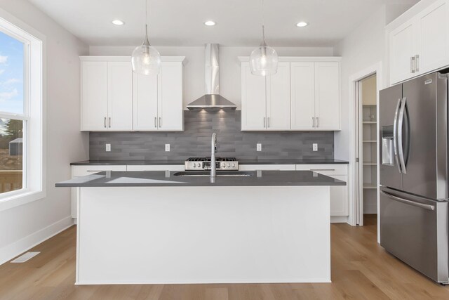 kitchen featuring a kitchen island with sink, stainless steel fridge, pendant lighting, and wall chimney range hood