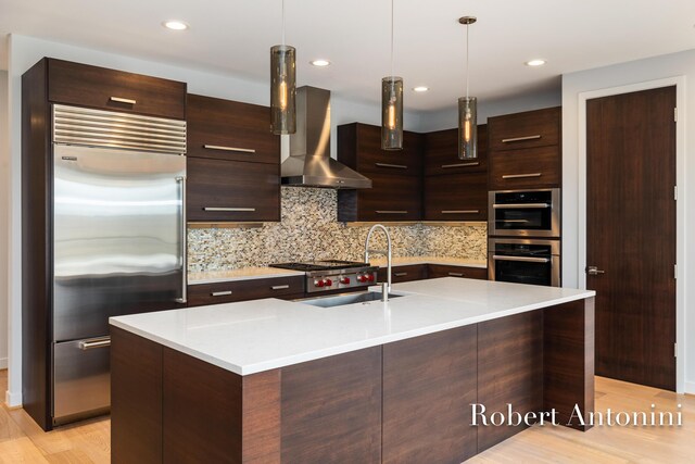 kitchen with a center island with sink, wall chimney exhaust hood, appliances with stainless steel finishes, decorative light fixtures, and light hardwood / wood-style floors