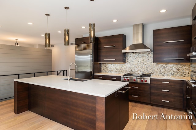 kitchen with sink, stainless steel appliances, wall chimney range hood, decorative light fixtures, and a kitchen island with sink