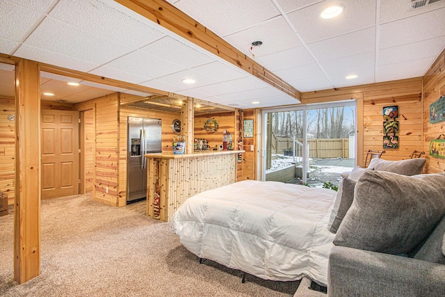 carpeted bedroom with a paneled ceiling, wood walls, stainless steel fridge with ice dispenser, and bar