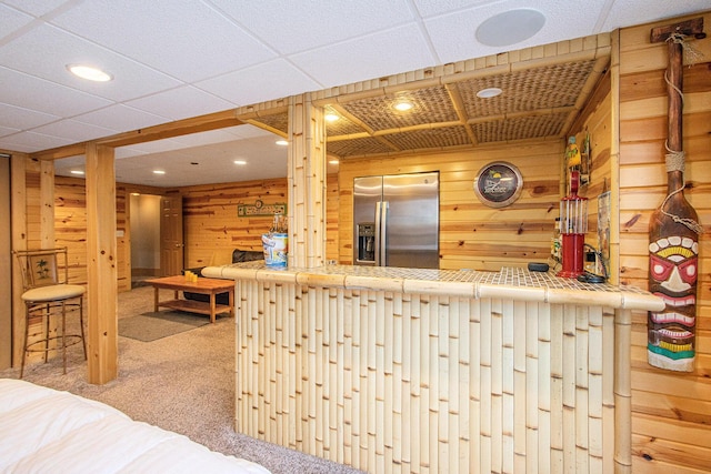 interior space with carpet flooring, wood walls, a drop ceiling, and stainless steel refrigerator with ice dispenser