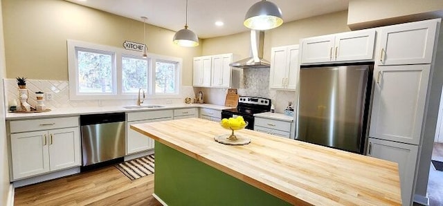 kitchen featuring appliances with stainless steel finishes, wall chimney exhaust hood, sink, decorative light fixtures, and butcher block counters