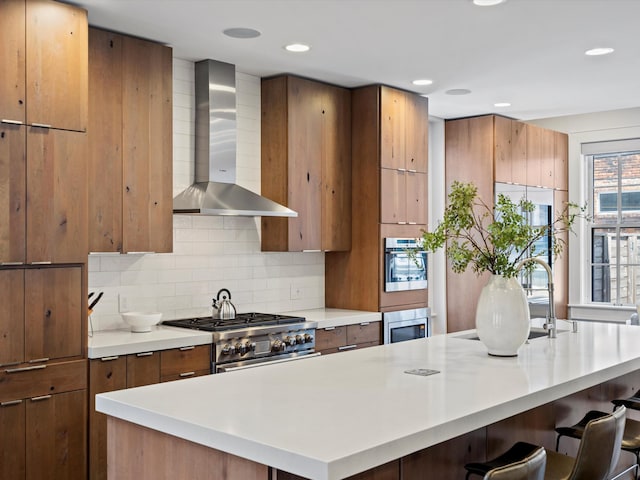 kitchen with wall chimney range hood, a kitchen breakfast bar, an island with sink, decorative backsplash, and appliances with stainless steel finishes