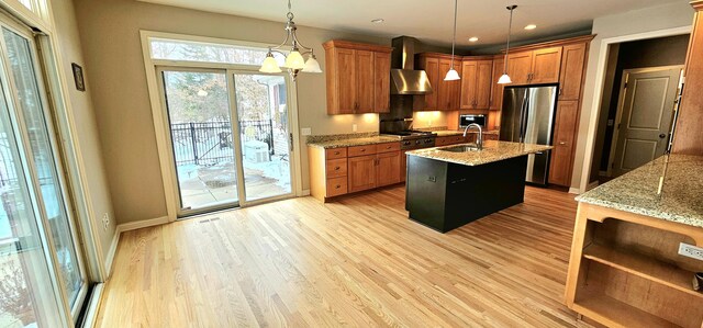 kitchen with wall chimney exhaust hood, stainless steel fridge, pendant lighting, a center island with sink, and light wood-type flooring