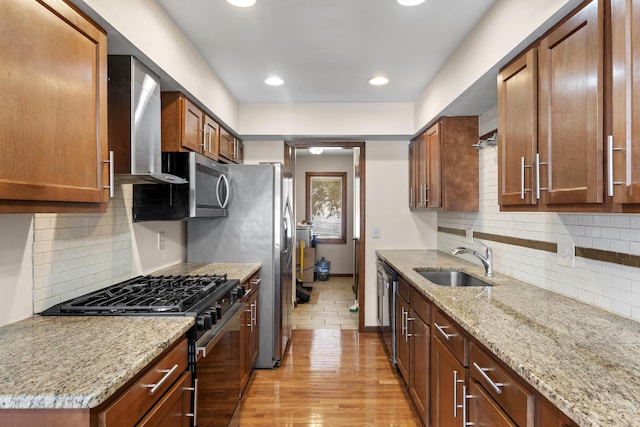 kitchen with sink, stainless steel appliances, light stone countertops, wall chimney range hood, and light hardwood / wood-style flooring