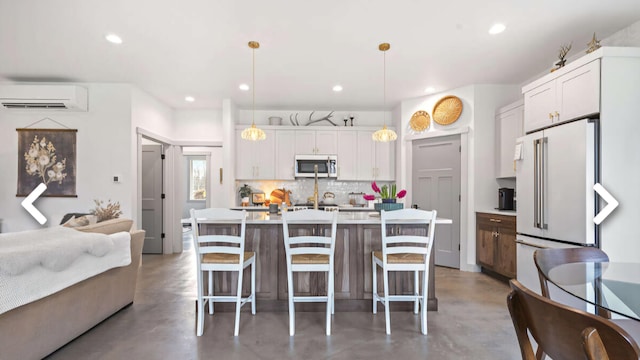 kitchen featuring high end fridge, a wall mounted AC, decorative light fixtures, white cabinetry, and an island with sink