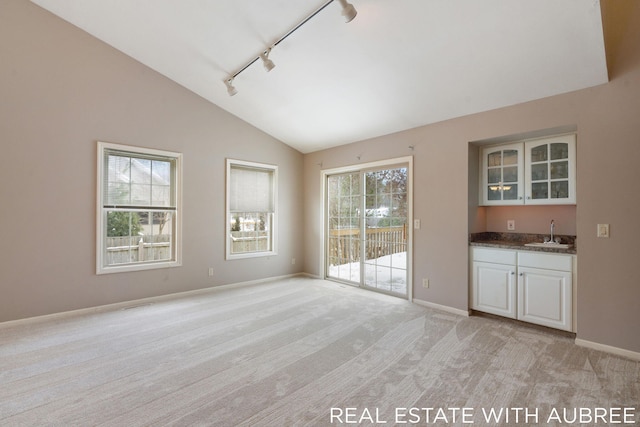 unfurnished living room with vaulted ceiling, track lighting, indoor wet bar, and light colored carpet