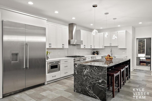 kitchen featuring light stone countertops, appliances with stainless steel finishes, wall chimney exhaust hood, a center island, and white cabinetry