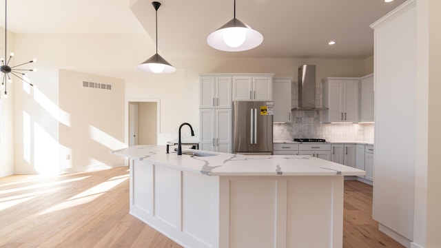 kitchen with stainless steel fridge, wall chimney exhaust hood, sink, hanging light fixtures, and an island with sink