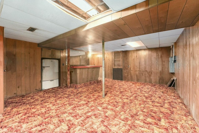 basement featuring carpet floors, white fridge, a drop ceiling, and wooden walls