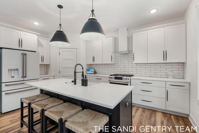 kitchen with wall chimney range hood, white refrigerator with ice dispenser, a center island with sink, high end stainless steel range, and white cabinets