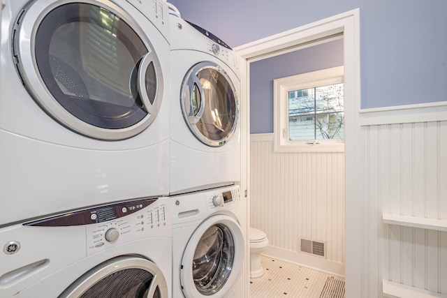 laundry area featuring a wainscoted wall, visible vents, stacked washing maching and dryer, tile patterned flooring, and laundry area