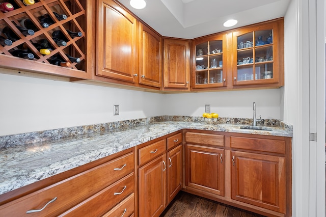 kitchen with light stone counters, dark wood-style flooring, a sink, brown cabinets, and glass insert cabinets