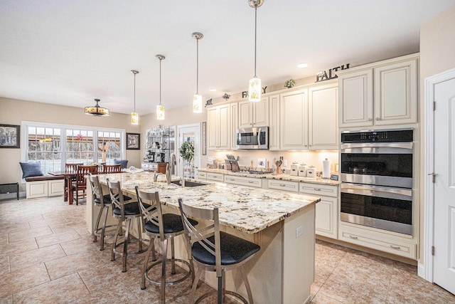 kitchen with a kitchen island with sink, cream cabinets, hanging light fixtures, sink, and appliances with stainless steel finishes