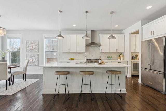 kitchen featuring wall chimney exhaust hood, high end refrigerator, pendant lighting, a kitchen island with sink, and white cabinets