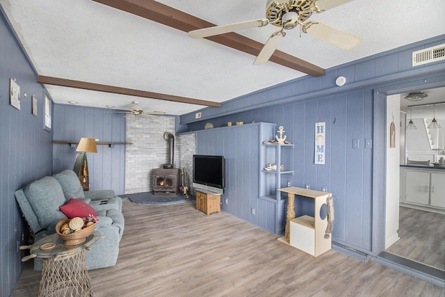 living room featuring beam ceiling, a wood stove, ceiling fan, wood-type flooring, and a textured ceiling