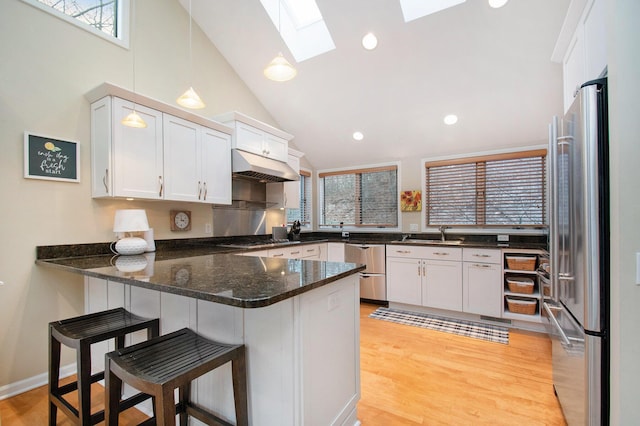 kitchen with kitchen peninsula, appliances with stainless steel finishes, a skylight, dark stone countertops, and white cabinetry