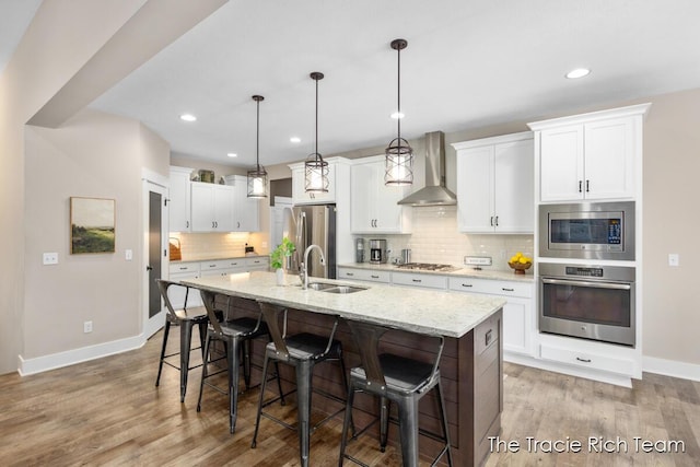 kitchen featuring wall chimney exhaust hood, white cabinetry, hanging light fixtures, appliances with stainless steel finishes, and an island with sink