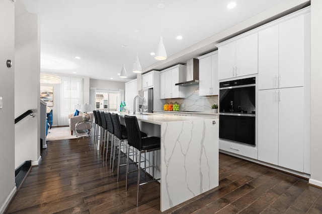 kitchen featuring a center island with sink, black double oven, wall chimney exhaust hood, white cabinetry, and a breakfast bar area