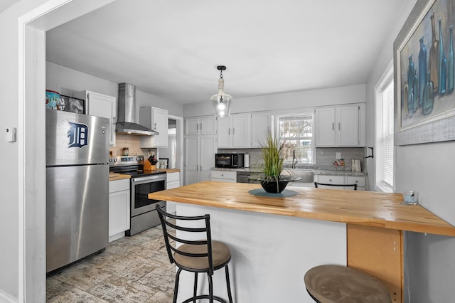 kitchen featuring white cabinetry, wall chimney exhaust hood, wooden counters, a kitchen bar, and appliances with stainless steel finishes