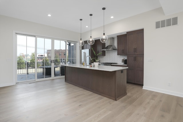 kitchen featuring backsplash, stainless steel appliances, a kitchen island with sink, wall chimney range hood, and hanging light fixtures