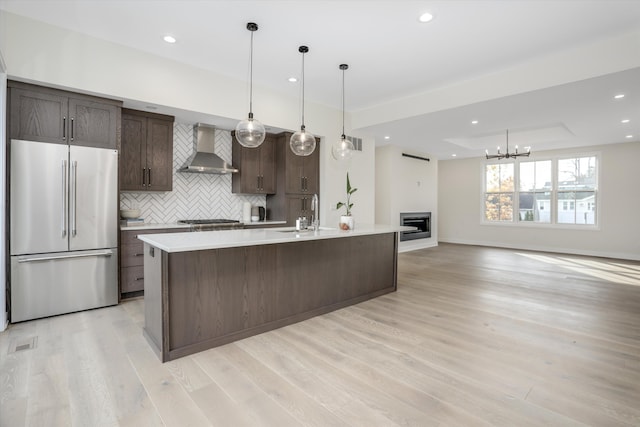 kitchen featuring dark brown cabinetry, wall chimney range hood, an island with sink, pendant lighting, and appliances with stainless steel finishes