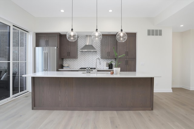 kitchen featuring wall chimney range hood, an island with sink, tasteful backsplash, high end fridge, and dark brown cabinetry