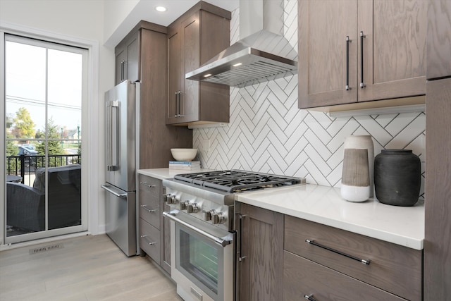 kitchen with wall chimney exhaust hood, high quality appliances, decorative backsplash, and light wood-type flooring