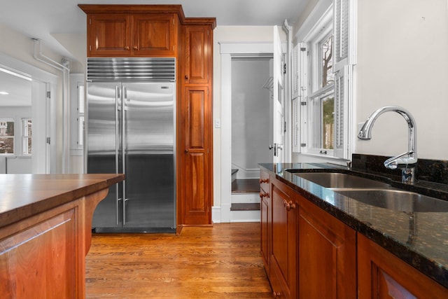 kitchen with dark stone countertops, sink, built in fridge, and light wood-type flooring