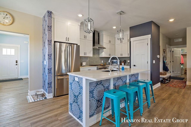 kitchen with stainless steel appliances, wall chimney range hood, decorative light fixtures, white cabinetry, and an island with sink