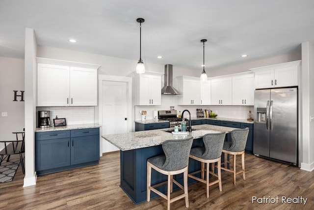 kitchen featuring pendant lighting, a kitchen island with sink, stainless steel appliances, white cabinets, and wall chimney exhaust hood