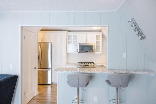 kitchen featuring white cabinetry, wood walls, appliances with stainless steel finishes, a kitchen breakfast bar, and light stone counters