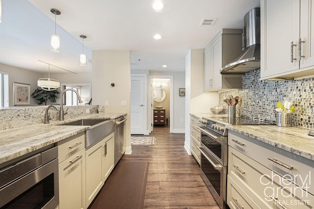 kitchen featuring light stone countertops, sink, wall chimney range hood, pendant lighting, and appliances with stainless steel finishes