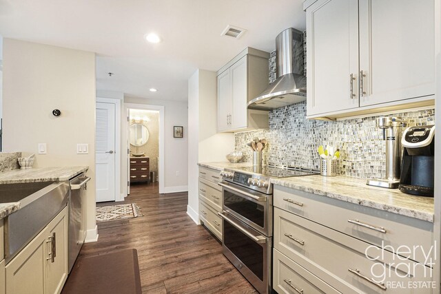 kitchen with backsplash, wall chimney range hood, dark hardwood / wood-style floors, light stone countertops, and stainless steel appliances