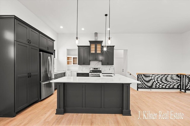 kitchen with an island with sink, sink, hanging light fixtures, stainless steel appliances, and light stone countertops