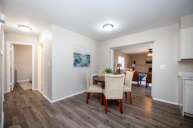 dining space with a brick fireplace, ceiling fan, and dark wood-type flooring