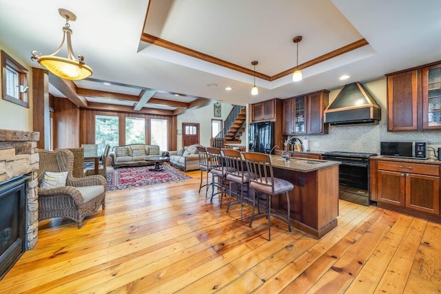 kitchen featuring a kitchen island with sink, black appliances, hanging light fixtures, premium range hood, and a tray ceiling