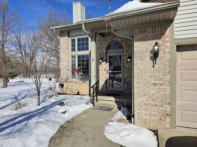 snow covered property entrance with a garage, brick siding, and a chimney