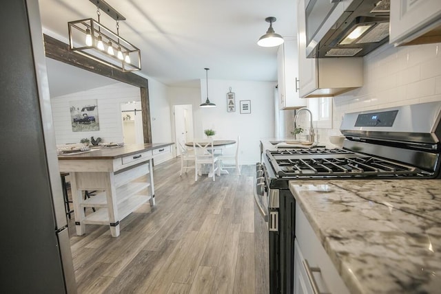 kitchen featuring wall chimney exhaust hood, light hardwood / wood-style flooring, hanging light fixtures, stainless steel gas stove, and white cabinets