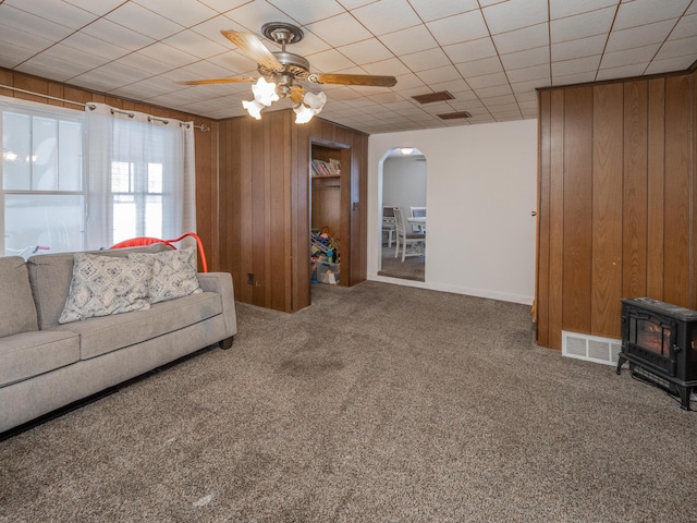 carpeted living room with ceiling fan, wooden walls, and a wood stove