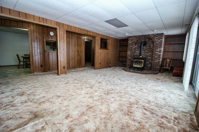 unfurnished living room with carpet floors, a wood stove, built in features, wood walls, and a paneled ceiling