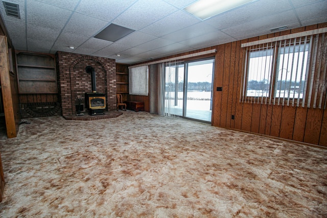 unfurnished living room featuring carpet floors, a wood stove, a water view, wood walls, and a paneled ceiling