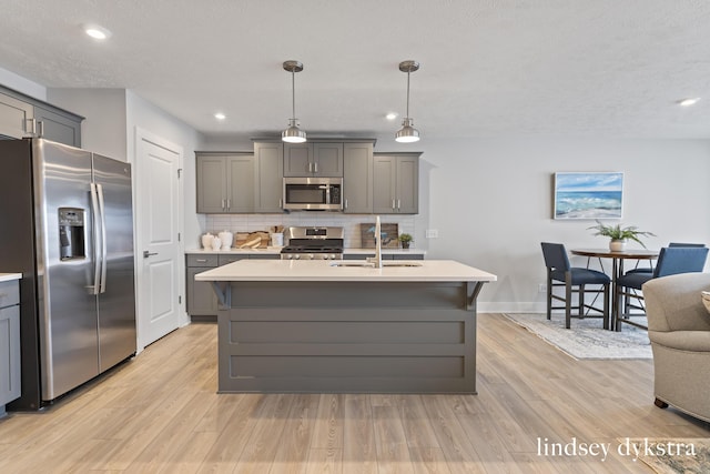 kitchen with a center island with sink, stainless steel appliances, gray cabinetry, tasteful backsplash, and hanging light fixtures