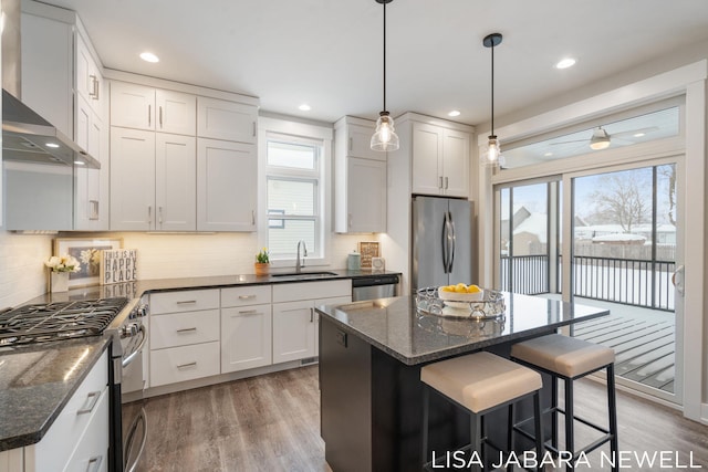 kitchen featuring wall chimney range hood, sink, appliances with stainless steel finishes, white cabinetry, and a center island