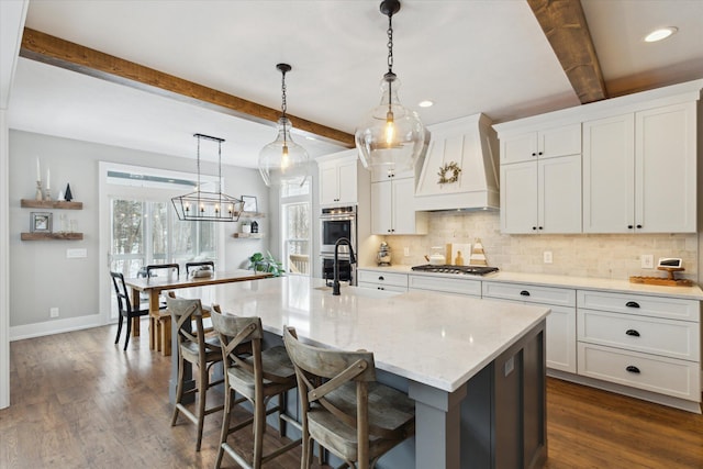 kitchen with custom exhaust hood, beamed ceiling, white cabinetry, stainless steel appliances, and a center island with sink