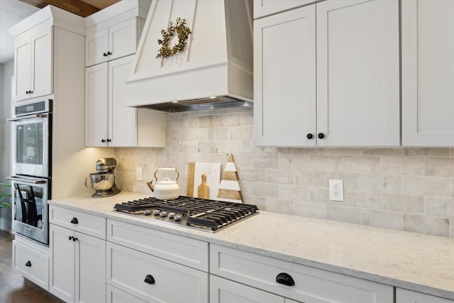 kitchen featuring light stone countertops, white cabinets, custom exhaust hood, stainless steel appliances, and backsplash