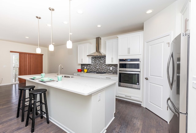 kitchen with sink, hanging light fixtures, wall chimney range hood, stainless steel appliances, and a center island with sink